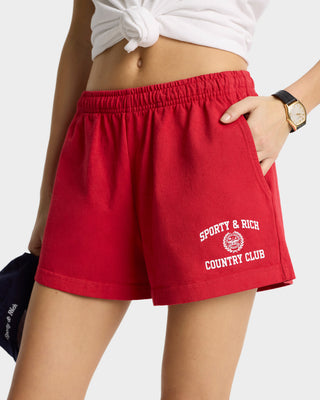 Person dressed in a white top and Varsity Crest Disco Short in Ruby/White, featuring an elastic waistband and Sporty & Rich Country Club text. A watch adorns the wrist as their hand rests casually in a pocket.