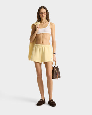 A woman in a white crop top, glasses, and the SR Runner Soft Disco Short in Almond/Navy with an elastic waistband holds a brown handbag against a plain background.