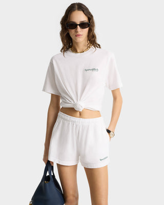 Woman in sunglasses wears a knotted white t-shirt and Serif Logo Disco Short in White/Forest with an elastic waistband, holding a navy bag.