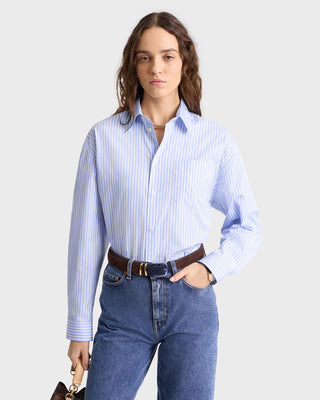 The woman in a SRC Oversized Shirt - Light Blue Striped with jeans holds a small handbag against a plain background.