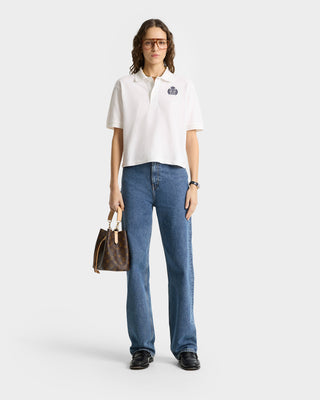 A woman in glasses wears an H&W Crest Pique Polo in White/Navy, blue jeans, black shoes, and carries a brown handbag.