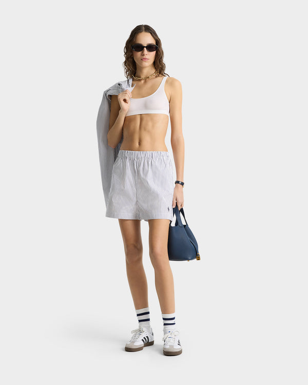 Woman in a white crop top, striped skirt, sneakers, blue bag and jacket, with sunglasses—her style pairs perfectly with the SRC Poplin Short - Blue Striped for a fresh unisex look.
