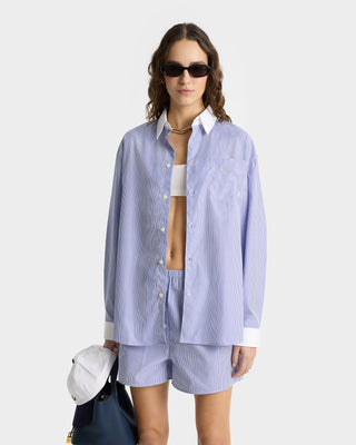 Woman in the SRC Shirt - Blue Striped, oversized fit, paired with matching shorts, white crop top, sunglasses, holding a navy bag and white cap.