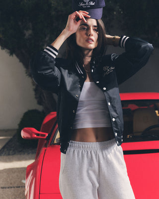 A woman in a black jacket and NY Health Club Sweatpant - Heather Gray/Navy, made of 100% cotton in Portugal, poses in front of a red sports car, adjusting her cap.