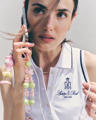 Woman wearing the Vendome Pique Polo Dress in White/Navy, holding a phone adorned with colorful beaded charms, hair blowing across her face.