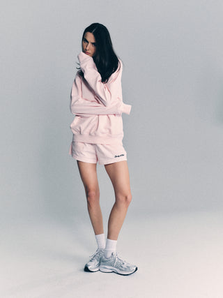 Someone wears the Syracuse Embroidered Disco short - Pink Dolphin/Navy, featuring heavy jersey fabric and an elastic waistband, paired with a pink sweatshirt, white socks, and silver sneakers. Made in Portugal.