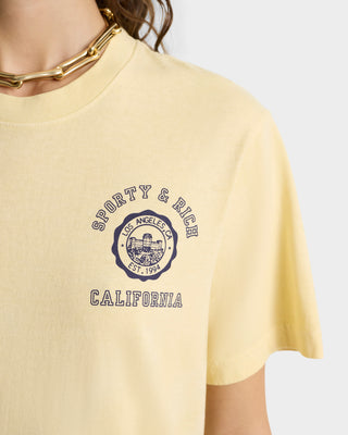 Person wearing the California Emblem T-Shirt in Almond/Navy by Sporty & Rich, made from 100% shrink-free cotton, paired with a chunky gold chain necklace.