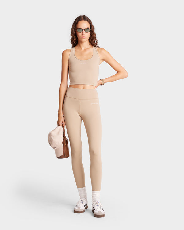 A person in the Serif Logo Legging in Elephant/White holds a cap, standing against a plain backdrop wearing chic sunglasses and sneakers.