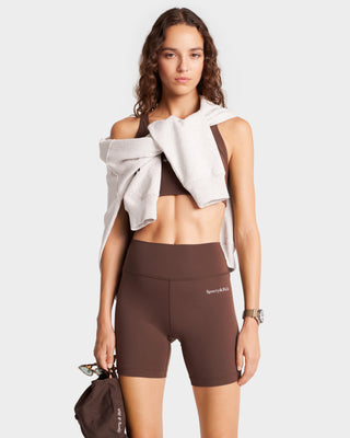 A woman exudes effortless style in Serif Logo Biker Shorts - Chocolate/White, with a sweatshirt draped over her shoulders. She holds a brown bag and wears a watch, perfectly blending fashion and function.