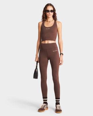 A woman stands confidently in luxury activewear, featuring Serif Logo Legging in Chocolate/White, sunglasses, a black bag, sneakers, and striped socks.