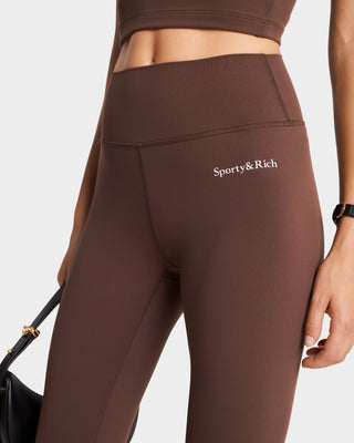 Close-up of a person wearing luxury activewear: Serif Logo Legging in Chocolate/White by Sporty & Rich, while holding a black bag.