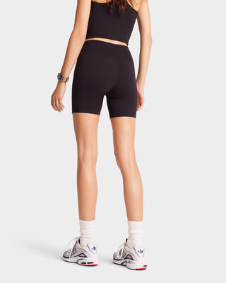 A person in high-waisted Serif Logo Biker Short - Black/White luxury activewear stands with their back to the camera, wearing sleek black sneakers against a pristine white background.