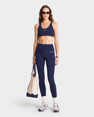 A person in Serif Logo Legging - Navy/White, paired with a stylish beige tote. They complete the look with sunglasses and sneakers, effortlessly blending comfort and style.