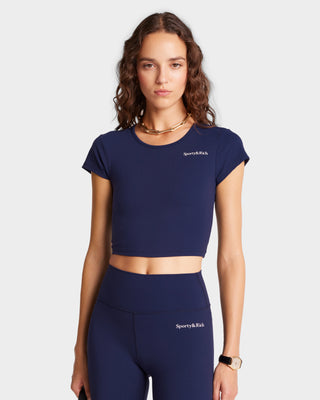 A woman in a luxury activewear ensemble featuring the Serif Logo Training Tee - Navy/White, paired with matching cropped leggings, accessorized with a gold necklace and watch.