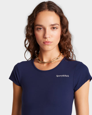 A person with curly hair wears the navy/white Serif Logo Training Tee by Sporty & Rich, paired with a gold chain necklace, set against a light background.