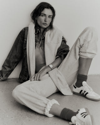 A person sits against a wall, wearing a denim jacket made from USA-grown cotton, paired with Serif Logo Sweatpant in Heather Gray/Navy and sneakers.