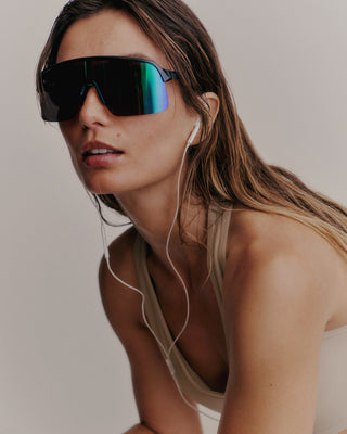 A person with long hair, wearing large sunglasses and earphones, showcases the Serif Logo V Neck Sports Bra in Elephant/White, glancing slightly to the side and exuding an air of luxury activewear.