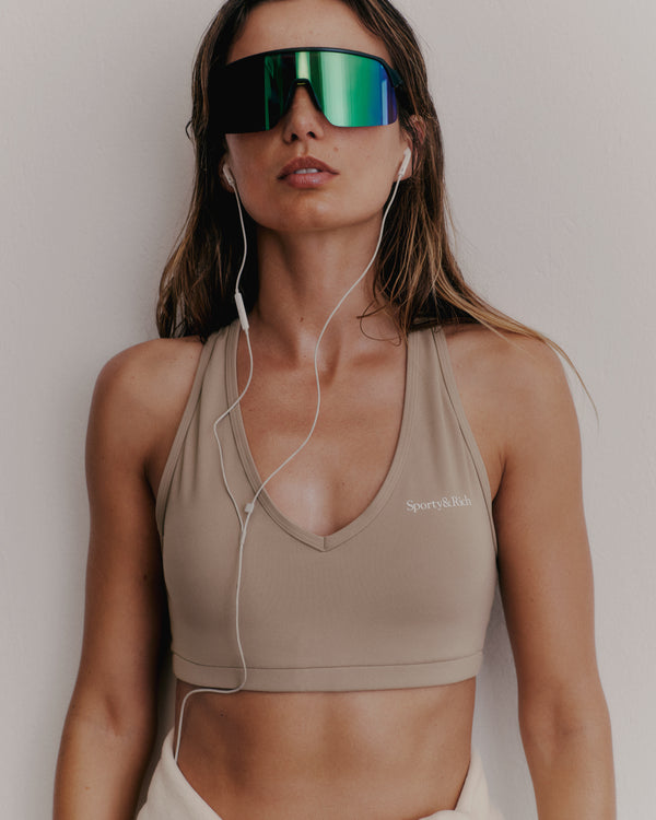 A woman in a Serif Logo V Neck Sports Bra in Elephant/White and large reflective sunglasses, wearing earphones, stands against a plain wall.