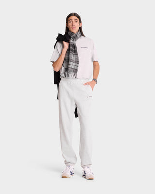 Wearing a Serif Logo Sweatpant in Heather Gray/Navy and a scarf, someone showcases unisex style against a white backdrop while holding a jacket. Their outfit features USA-grown cotton and is proudly made in the USA.