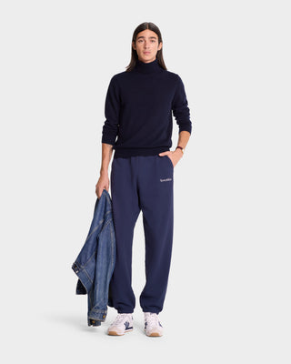 A person in a Serif Logo Sweatpant (Navy/White) and a matching turtleneck, made from premium USA cotton, holds a denim jacket and wears white sneakers against a plain background.