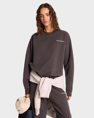 A person in a Serif Logo Crewneck sweatshirt in Faded Black/White and pants, made from unisex USA-grown cotton featuring the Sporty & Rich logo, holds a cap with a light jacket tied at their waist.