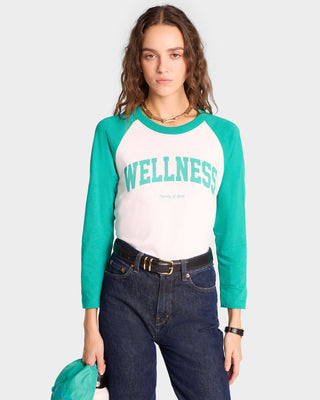 Woman in a Wellness Ivy Baseball Tee - White/Spring Green, made from USA grown cotton, and jeans, standing against a plain background and looking at the camera.
