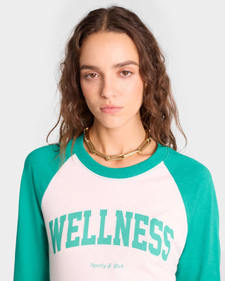 Woman with wavy brown hair wears the Wellness Ivy Baseball Tee in White/Spring Green, made from USA-grown cotton, paired with a chunky gold necklace.