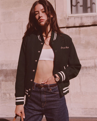 A woman in casual wear, sporting the Syracuse Wool Varsity Jacket - Forest with embroidered graphics, stands against a sunlit wall.
