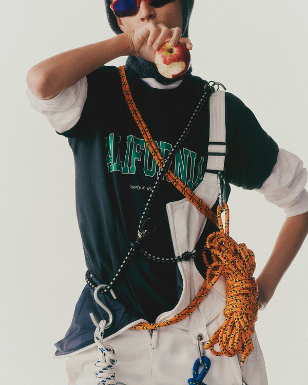 A person in a shrink-free, 100% cotton California T-Shirt - Faded Black/Spring Green, wearing a harness and ropes, eating an apple while sporting sunglasses and a beanie.