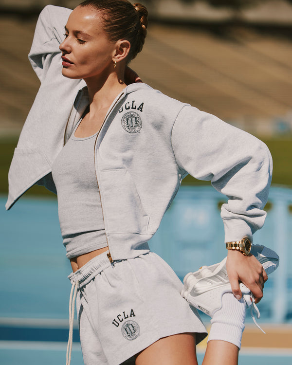 model wearing Heather grey full-zip hoodie with navy 'UCLA' text and the official UCLA seal on the left chest, featuring front pockets, a hood, and small 'Sporty&Rich' text on the left sleeve.