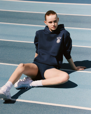 model wearing navy hoodie sweatshirt with white 'UCLA Bruins' text on the left chest and small 'Sporty&Rich' text on the left sleeve cuff, featuring ribbed hem and cuffs.