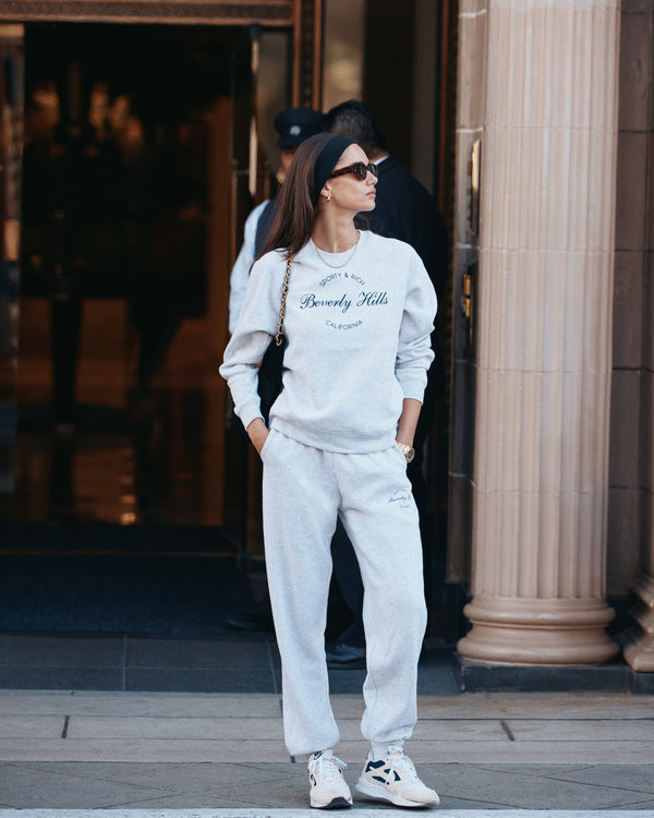 A woman in a Beverly Script Soft Crewneck - Heather Gray/Navy and sunglasses stands confidently outside a building, her hands tucked into the crewnecks pockets.