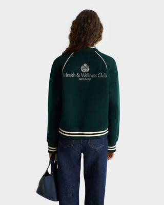 Individual wearing the H&W Crest Double Face Varsity Jacket in Forest/White with Health & Wellness Club on the back, holding a blue bag and facing away.