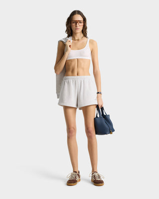 Person in sportswear wearing SR Runner Soft Disco Short in heather gray, holding a jacket and blue bag, while wearing glasses and brown sneakers with an elastic waistband against a plain background.