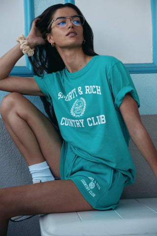 Woman in teal Sporty & Rich Country Club T-shirt and Varsity Crest Soft Disco Short in spring green/white with an elastic waistband, sitting with one knee up against a wall.