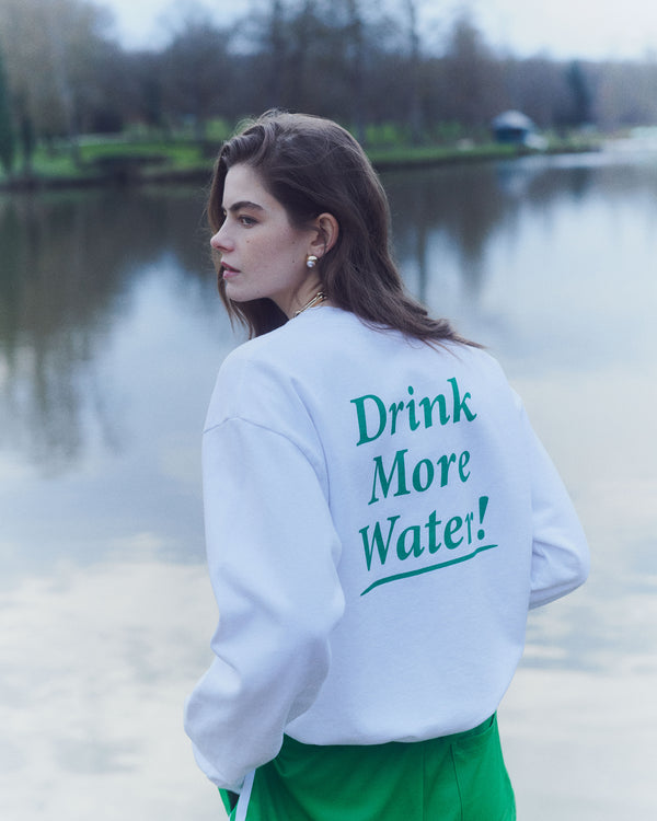 A woman by a lake wears the Drink More Water Crewneck - White/Verde, featuring Drink More Water! in green on the back. Made in the USA from 100% cotton, its pre-shrunk for comfort and quality.
