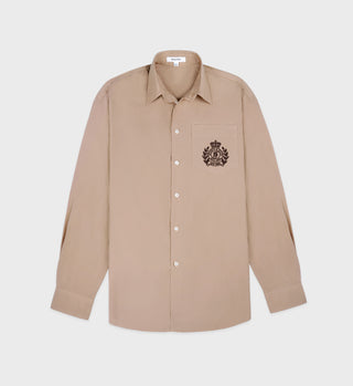 The H&W Crest Oversized Shirt in light tan/chocolate, made of cotton, features a black embroidered crest on its chest pocket and is laid flat on a white background.