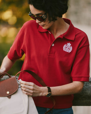Woman in H&W Crest Pique Polo - Ruby/White with sunglasses and watch, holding a beige bag outdoors.