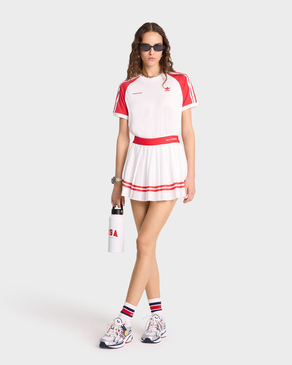 Woman wearing the Serif Logo Pleated Skirt in White/Sports Red, paired with a matching tennis outfit, sunglasses, sneakers, and holding a water bottle.