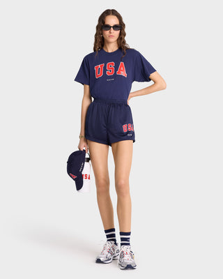 Woman in the USA T-Shirt - Navy/Sports Red/White made from USA grown cotton, paired with shorts, holding a cap and bottle, wearing sneakers and sunglasses.