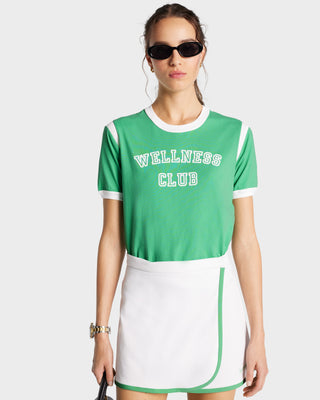 Woman in sunglasses wears the Wellness Club Sports Tee - Verde/White, made of soft cotton and garment dyed, paired with a white tennis skirt against a plain background.