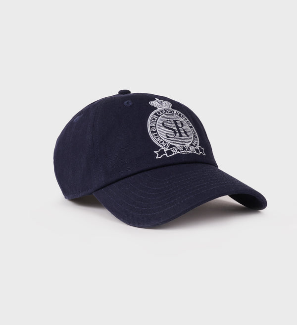 The Royal Club Hat - Navy/White is a navy blue cotton twill dad hat featuring an embroidered white crest and logo on the front, viewed at an angle. It includes an adjustable closure for a comfortable fit.