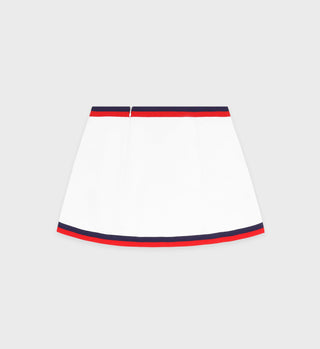 The Serif Logo Mila Skirt in white, navy, and fire red is a mini trapeze skirt made of stretch technical jersey with red and navy stripes on the waistband and hem, shown against a plain background.