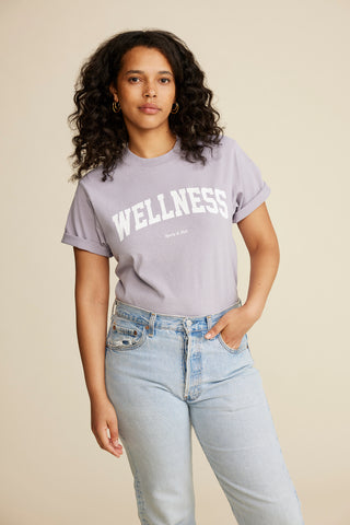 A woman with curly hair wears the Wellness Ivy T-Shirt in Faded Lilac/White, made from 100% USA-grown cotton and crafted in Los Angeles, paired with light blue jeans against a beige background.