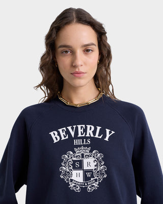 A woman stands against a plain background wearing the navy Beverly Hills Crest Soft Crewneck, which features a crest design and raglan sleeves.