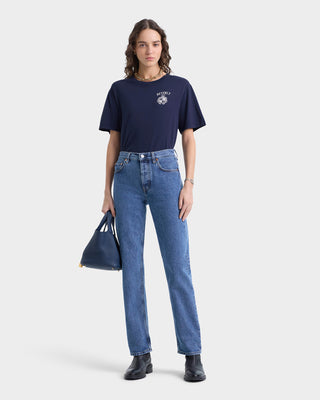 In the Beverly Hills Crest Soft T-Shirt - Navy/White and jeans, a person stands against a light background, holding a dark bag. The pre-shrunk shirt ensures lasting comfort and style.