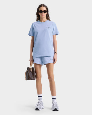 Clad in a Wimbledon T-Shirt - Washed Hydrangea/Navy, with sunglasses on, carrying a brown bag, and sporting white sneakers against a plain backdrop.