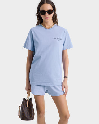 A woman wearing a Wimbledon T-Shirt in Washed Hydrangea/Navy and shorts, made from USA-grown cotton, is accessorized with sunglasses and a brown handbag against a plain background.