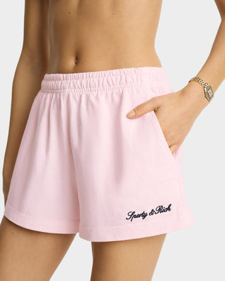 A person wearing Syracuse Embroidered Disco short in Pink Dolphin/Navy with an elastic waistband, Sporty & Rich logo on the hem, and a hand in the pocket.