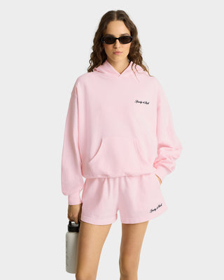 A woman wears the Syracuse Embroidered Hoodie - Pink Dolphin, made from 100% USA-grown cotton with an embroidered logo and pink color, styled with shorts and sunglasses while holding a water bottle against a plain background.